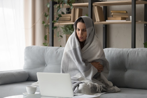 A woman sitting on a couch with a blanket with a laptop in front of her.