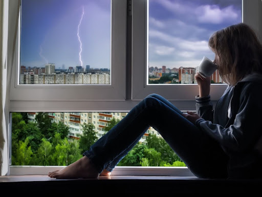 A woman sitting near a window drinking a cup of coffee with lightning and storm clouds in the sky.